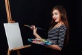 Cute beautiful girl artist painting a picture on canvas an easel. Space for text. Studio black background. Royalty Free Stock Photo