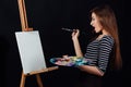 Cute beautiful girl artist painting a picture on canvas an easel. Space for text. Studio black background. Royalty Free Stock Photo