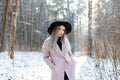 Cute beautiful young woman with blond hair in a vintage chic black hat in a pink elegant coat posing in a winter park Royalty Free Stock Photo