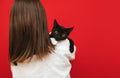 Cute black cat lies on the shoulders of a woman in a white shirt and looks into the camera on a red background. Handsome pet in a Royalty Free Stock Photo