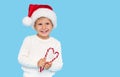 A cute blond baby boy in a Santa Claus hat holding a heart-shaped lollipop cane on a blue background. Royalty Free Stock Photo