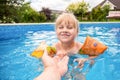 A cute blond baby girl swims at swimmig pool with blue colored water outdoors, smiles and hold parent`s hand.