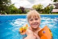 A cute blond baby girl swims at swimmig pool with blue colored water outdoors, smiles and hold parent`s hand.