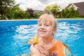 A cute blond baby girl swims at swimmig pool with blue colored water outdoors, smiles and hold parent`s hand.