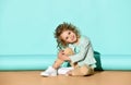 Cute blond girl in casual clothing sits on floor hugging knee, tilting her head and smiling. portrait on turquoise background Royalty Free Stock Photo