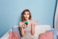 Cute blond girl with curly hair sitting on a sofa and holding a blue cupcake. Colorful pillows on the sofa. Girl looks Royalty Free Stock Photo