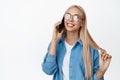 Cute blond girl in glasses flirting and talking on mobile phone, having a call, standing over white background Royalty Free Stock Photo