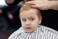 Cute blond smiling baby boy with blue eyes in a barber shop having haircut by hairdresser. Hands of stylist with tools. Royalty Free Stock Photo