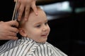 Cute blond smiling baby boy with blue eyes in a barber shop having haircut by hairdresser. Hands of stylist with tools. Children`s Royalty Free Stock Photo