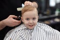 Cute blond smiling baby boy with blue eyes in a barber shop having haircut by hairdresser. Hands of stylist with tools. Children`s