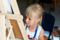 Cute blond smiling caucasian kid painting on wooden easel in class workshop lesson at art studio. Little girl holding brush in