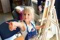 Cute blond smiling caucasian kid painting on wooden easel in class workshop lesson at art studio. Little girl holding brush in Royalty Free Stock Photo