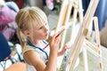 Cute blond smiling girl painting on easel in workshop lesson at art studio. Kid holding brush in hand and having fun drawing with