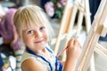 Cute blond smiling girl painting on easel in workshop lesson at art studio. Kid holding brush in hand and having fun drawing with