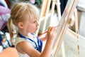 Cute blond smiling girl painting on easel in workshop lesson at art studio. Kid holding brush in hand and having fun