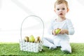 Baby holding yellow chicken egg while sitting on green grass near straw basket with Easter eggs isolated on white Royalty Free Stock Photo