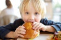 Cute blonde boy eating large hamburger at fastfood restaurant. Unhealthy meal for kids. Junk food Royalty Free Stock Photo