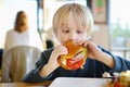 Cute blonde boy eating large hamburger at fastfood restaurant. Unhealthy meal for kids. Junk food