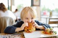 Cute blonde boy eating large hamburger at fastfood restaurant. Unhealthy meal for kids. Junk food