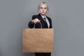 Cute blonde businesswoman in formal suit holding empty blank brown paper shopping bag on grey background Royalty Free Stock Photo