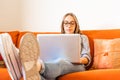 Cute blonde girl with eyeglasses sitting at home sofa focused on study or work at laptop computer with feet resting on table.