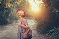 Cute blonde girl with short haircut is walking with bag in forest on sunset background. She wears hat, sunglasses Royalty Free Stock Photo