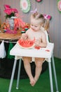 A cute blonde girl sits in a children`s white chair and eats a slice of a juicy watermelon in summer Royalty Free Stock Photo