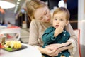 Cute blonde little boy eating in a fast food courte cafe or restaurant with his young mother during shopping in a mall. Healthy Royalty Free Stock Photo