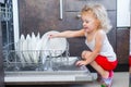 Cute blonde toddler girl helping in the kitchen taking plates out of dish washing machine Royalty Free Stock Photo