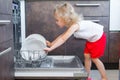 Cute blonde toddler girl helping in the kitchen taking plates out of dish washing machine Royalty Free Stock Photo