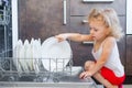 Cute blonde toddler girl helping in the kitchen taking plates out of dish washing machine Royalty Free Stock Photo