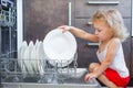 Cute blonde toddler girl helping in the kitchen taking plates out of dish washing machine Royalty Free Stock Photo
