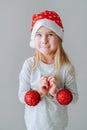 Cute blonde toddler girl wearing a santa hat holding two red Christmas balls of Christmas tree decoration against