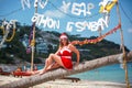 Cute blonde woman in red dress, sunglasses and santa hat sitting on palm tree at exotic tropical beach. Holiday concept Royalty Free Stock Photo