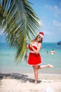 Cute blonde woman in red dress, sunglasses and santa hat stands at palm tree on exotic tropical beach. Holiday concept Royalty Free Stock Photo