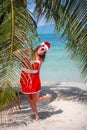 Cute blonde woman in red dress, sunglasses and santa hat stands at palm tree on exotic tropical beach. Holiday concept Royalty Free Stock Photo