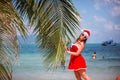 Cute blonde woman in red dress, sunglasses and santa hat stands at palm tree on exotic tropical beach. Holiday concept Royalty Free Stock Photo