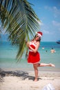 Cute blonde woman in red dress, sunglasses and santa hat stands at palm tree on exotic tropical beach. Holiday concept Royalty Free Stock Photo