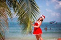 Cute blonde woman in red dress, sunglasses and santa hat stands at palm tree on exotic tropical beach. Holiday concept Royalty Free Stock Photo