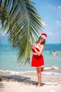 Cute blonde woman in red dress, sunglasses and santa hat stands at palm tree on exotic tropical beach. Holiday concept Royalty Free Stock Photo