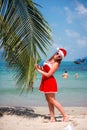 Cute blonde woman in red dress, sunglasses and santa hat stands at palm tree on exotic tropical beach. Holiday concept Royalty Free Stock Photo