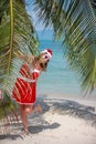 Cute blonde woman in red dress, sunglasses and santa hat stands at palm tree on exotic tropical beach. Holiday concept Royalty Free Stock Photo