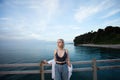 Cute blonde woman in white shirt and tank top standing on pier on the background of sea, cloudy sky and coastline with green trees