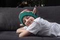 Cute boy in christmas elf hat is resting on the sofa with closed eyes on the background of Christmas tree. Anticipation of holiday