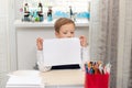 A cute boy first grader in a school uniform at home while at his desk holds a sheet of blank paper.