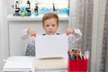 A cute boy first grader in a school uniform at home while isolated at his desk holds a sheet of blank paper.