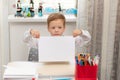 A cute boy first grader in a school uniform at home while isolated at his desk holds a sheet of blank paper.