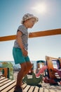 A cute boy in a hat walks on a wooden bridge to the beach. A playground, sea and sun rays in the background Royalty Free Stock Photo