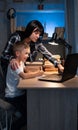 Cute boy with mother doing homework at home using laptop and books at night, online schooling Royalty Free Stock Photo