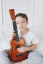 Boy playing a small guitar sitting on the bed Royalty Free Stock Photo